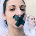 photo of a young woman outside at a Chain-In protest. She wears a white veil and her dark hair is tied back. She has a black "X" taped over her mouth. Another woman stands behind her with a sign that reads "Stop child marriage in the U.S."