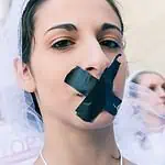 photo of a young woman outside at a Chain-In protest. She wears a white veil and her dark hair is tied back. She has a black "X" taped over her mouth. Another woman stands behind her with a sign that reads "Stop child marriage in the U.S."