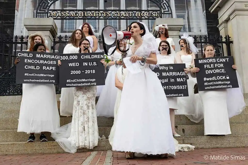 Photo by Matilde Simas of Capture Humanity: Fraidy Reiss speaks to the crowd via a megaphone during the September 22 Chain-In in front of the Massachusetts state house. Several protesters are on the stairs behind her, all in white bridal gowns and veils with chains on their wrists and a black "x" taped over their mouths. The four in front hold signs.