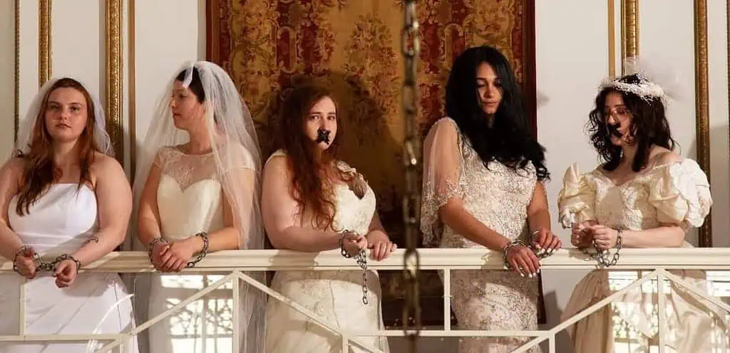 a group of women in bridal gowns and veils stand along a railing inside a beautiful mansion. They look sad, and many have chains around their wrists and tape over their mouths. A chain hangs through the center of the photo
